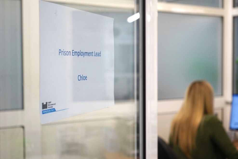 A view through a doorway, the door is ajar and has a notice "Prison Employment Lead." There is a female in the background with blonde hair and her back to the door.