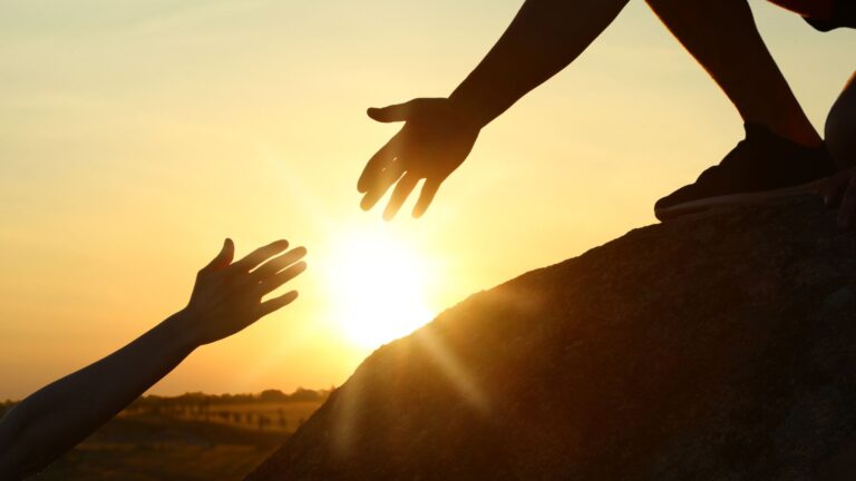 Photo of a person reaching out for another person's hand in front of a sunset