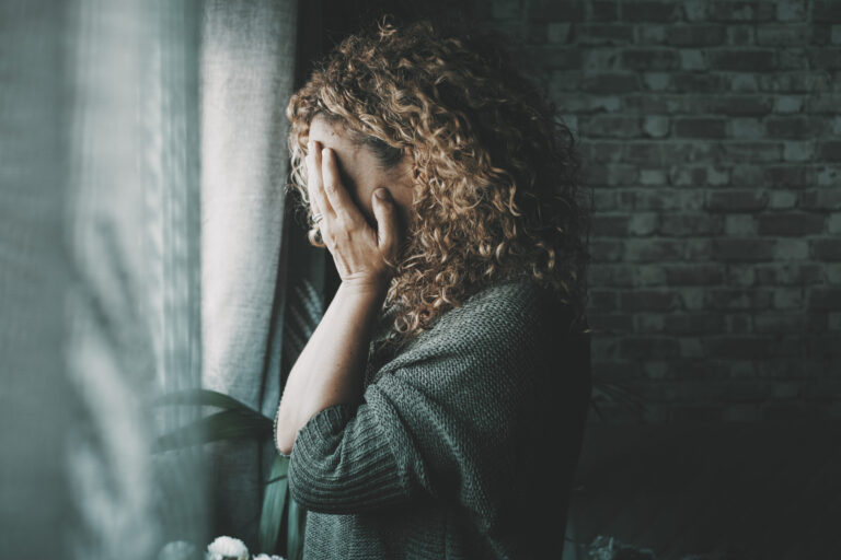 A photo of a female in a dark room, standing in front of a window with her head in her hands