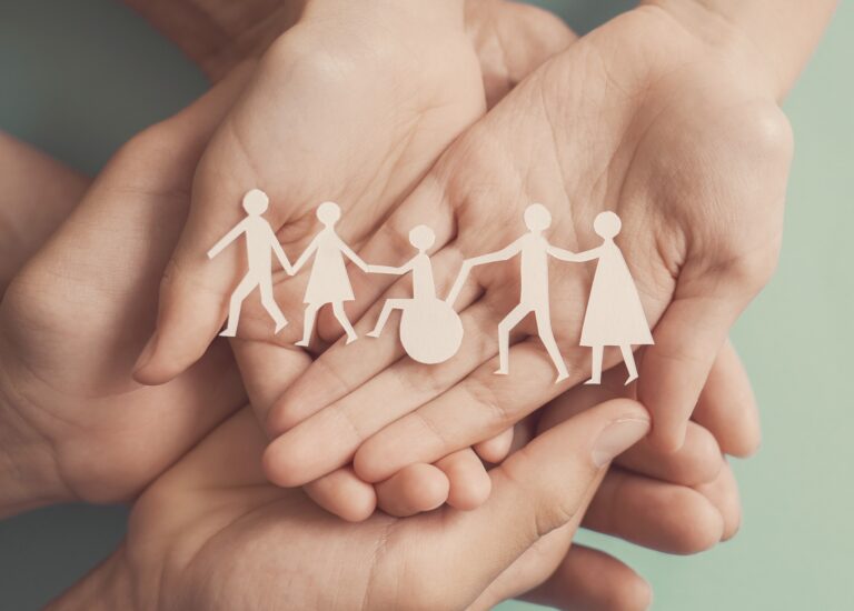 A conceptual photo photo of an adult and child's hands holding a paper cutout of different shaped people including a person in a wheelchair