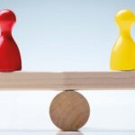 An image of a toy wooden seesaw which is balanced with a yellow chess pawn on one side and a red on the other.