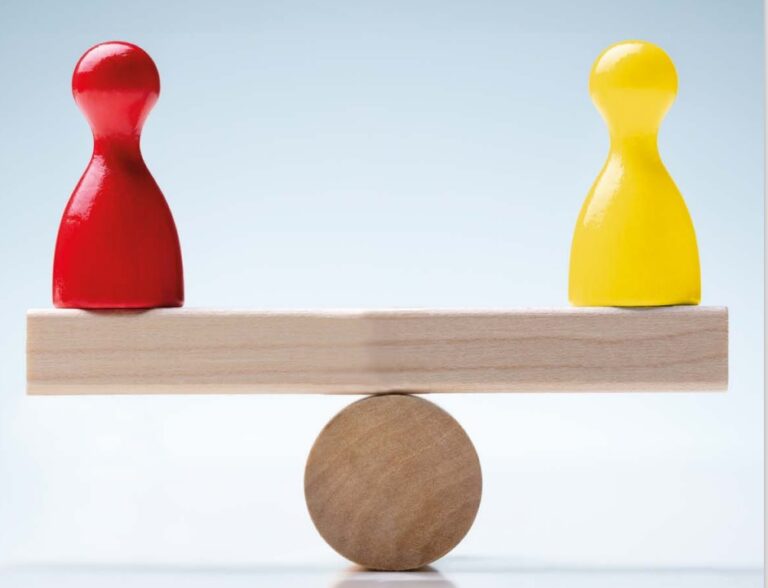 An image of a toy wooden seesaw which is balanced with a yellow chess pawn on one side and a red on the other.