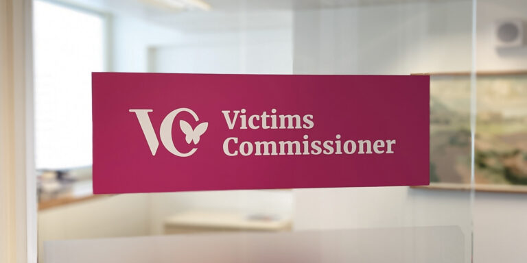 A sign on a glass door with 'VC - Victims' Commissioner' written on it. The logo is pink with white text.