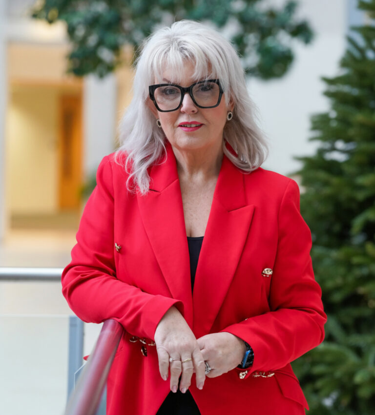 A portrait of Baroness Newlove, dressed in a vibrant red blazer with gold accents. She is leaning on a railing in an indoor setting.