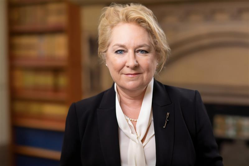 Headshot of the Lady Chief Justice smiling warmly at the camera, wearing a blazer and blouse in front of a bookcase and an ornate fireplace.